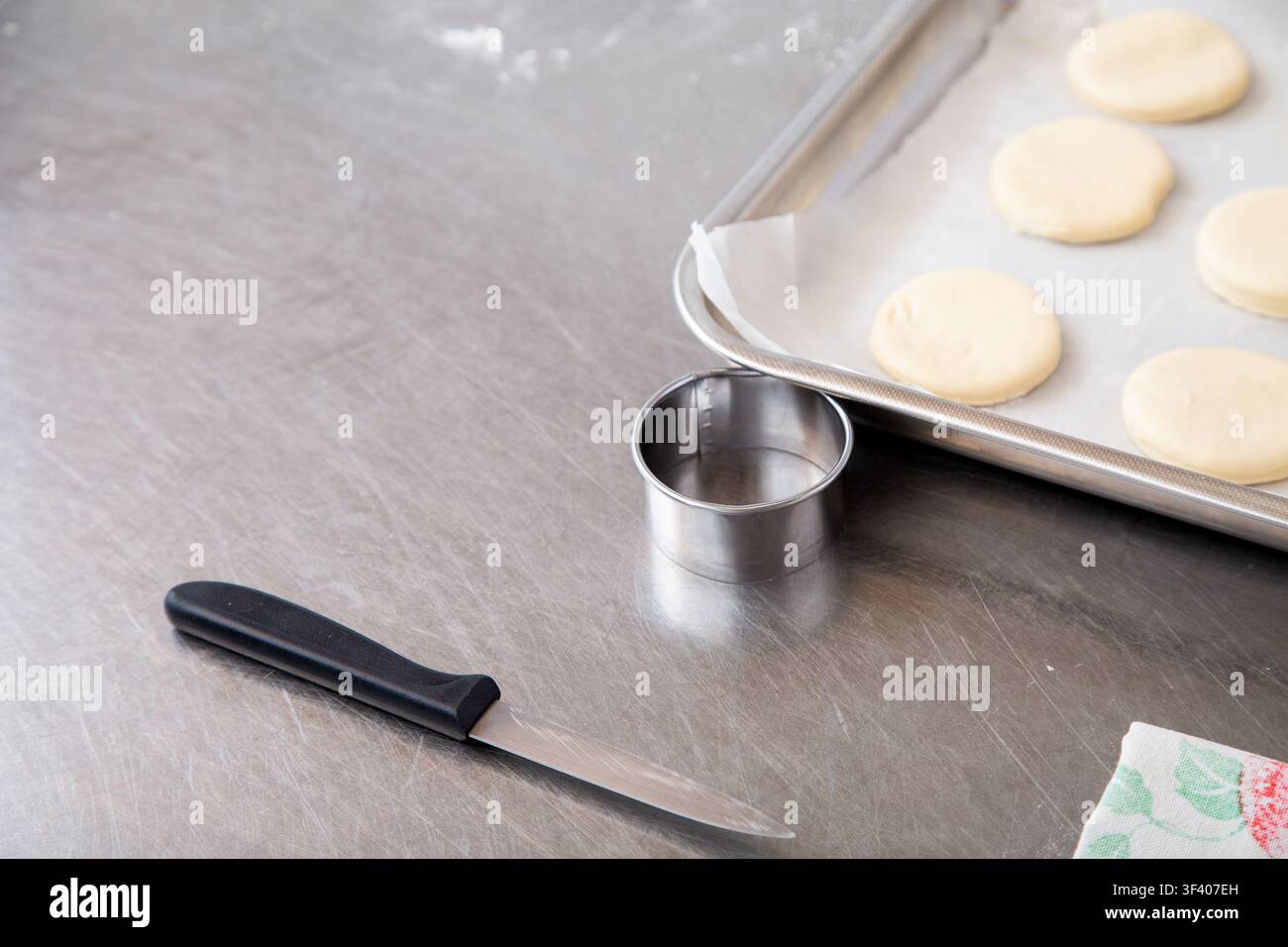 Teigringe auf backpapier ausgelegtem Backblech neben Messer und Metallring auf gebürsteter Edelstahl-Küchenarbeitsplatte, bereit zum Backen Stockfoto