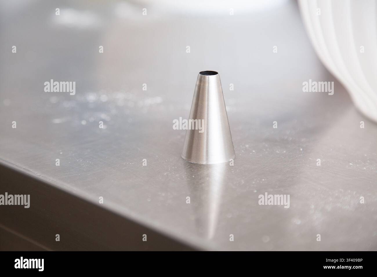 Minimalistischer Edelstahlkegel steht auf gebürsteter Metalloberfläche mit weichen hellen Reflexionen und geringer Schärfentiefe, wodurch eine saubere Industrieästhetik entsteht Stockfoto