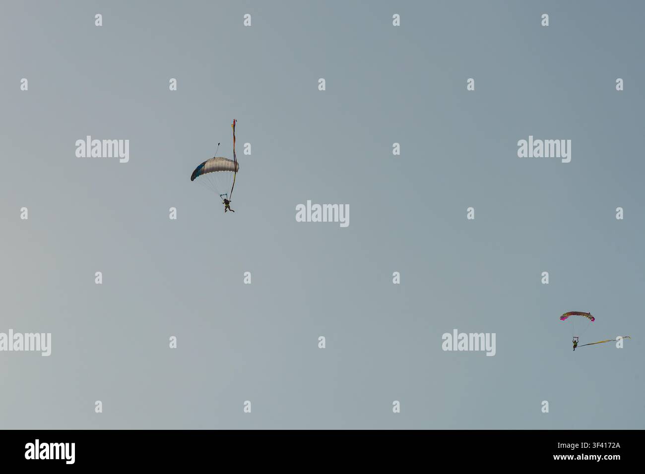 Zwei Fallschirmspringer gleiten mit rechteckigen Fallschirmen abwärts, während sie lange bunte Bänder gegen einen klaren Himmel hinter sich herziehen. Stockfoto