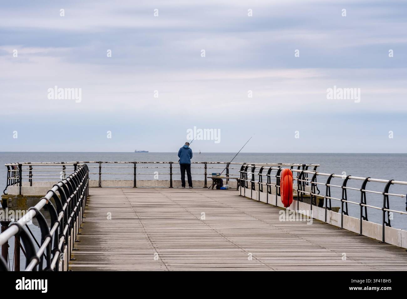 Ein einsamer Angler steht am Ende des Saltburn-Piers und wirft in die ruhigen Sommergewässer, während das Abendlicht über der Nordsee weicher wird. Stockfoto