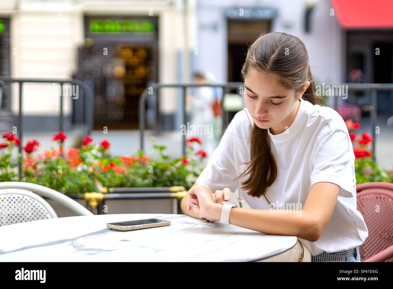 Junge Frau überprüft Smartwatch in einem Straßencafé. Junge Frau überprüft ein Smartwatch, während sie an einem Tisch im Straßencafé sitzt, mit einem Smartphone in der Nähe und Stockfoto