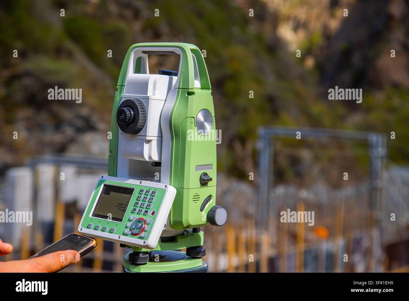 Nahaufnahme einer grün-weißen Totalstation auf einem Stativ im Freien. Eine Hand hält ein Smartphone in der Nähe des Displays des Geräts, was auf Datenübertragung hinweist Stockfoto