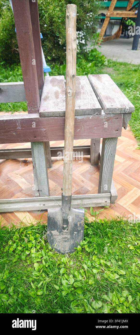 Alte Gartenschaufel mit trockener Erde auf der Metallklinge, die an einer Holzbank im Freien lehnt Stockfoto