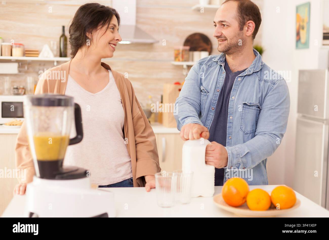 Fröhliches Paar macht einen Smoothie mit gemischten Früchten in einem Mixer, bereitet ein frisches hausgemachtes Getränk in der Küche zu für frische Zutaten und alltäglichen Küchenlebensstil-Kontext. Stockfoto