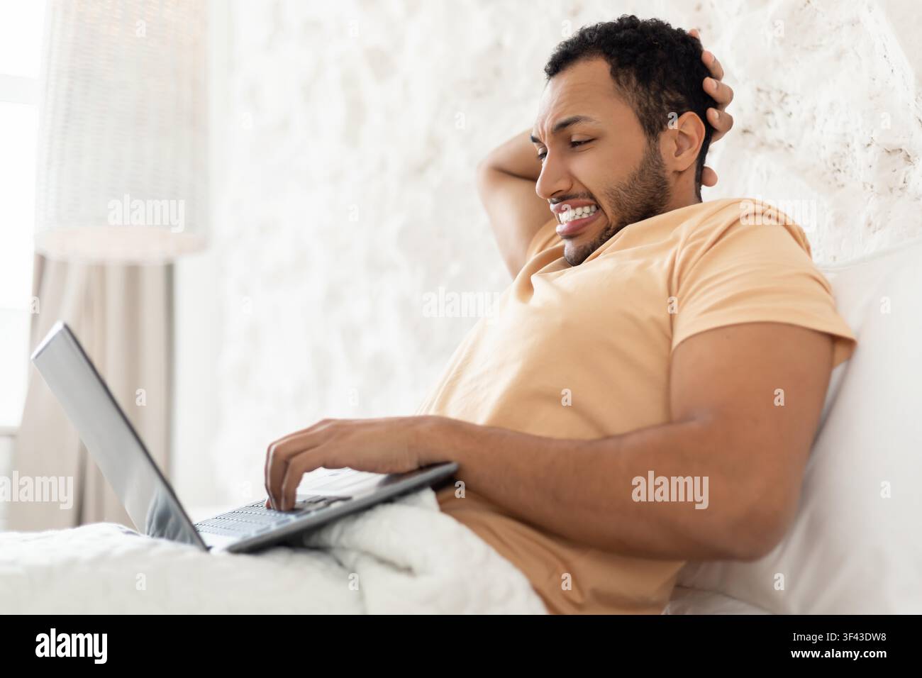 Glücklicher schwarzer Mann arbeitet an einem Laptop, während er im Bett entspannt, Home-Office-Konzept Stockfoto