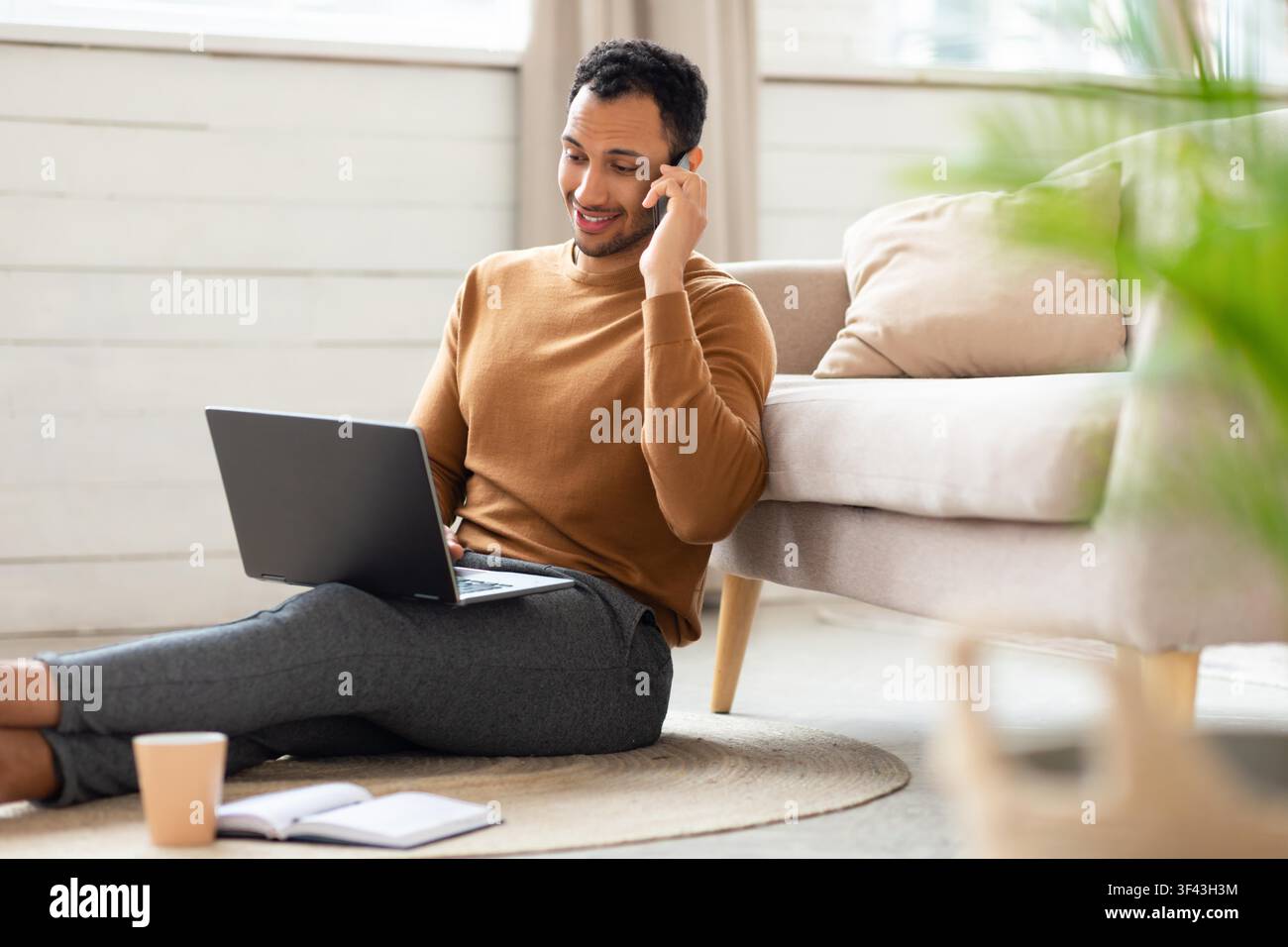 Lächelnder schwarzer Mann arbeitet an einem Laptop und telefoniert zu Hause, Geschäftskonzept Stockfoto