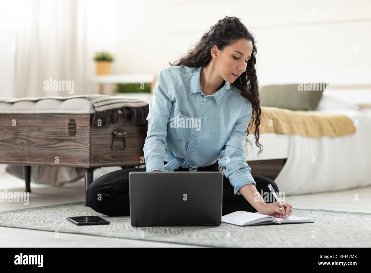 Konzentrierte junge Frau gemischter Herkunft arbeitet an einem Laptop und macht Notizen im Homeoffice Stockfoto