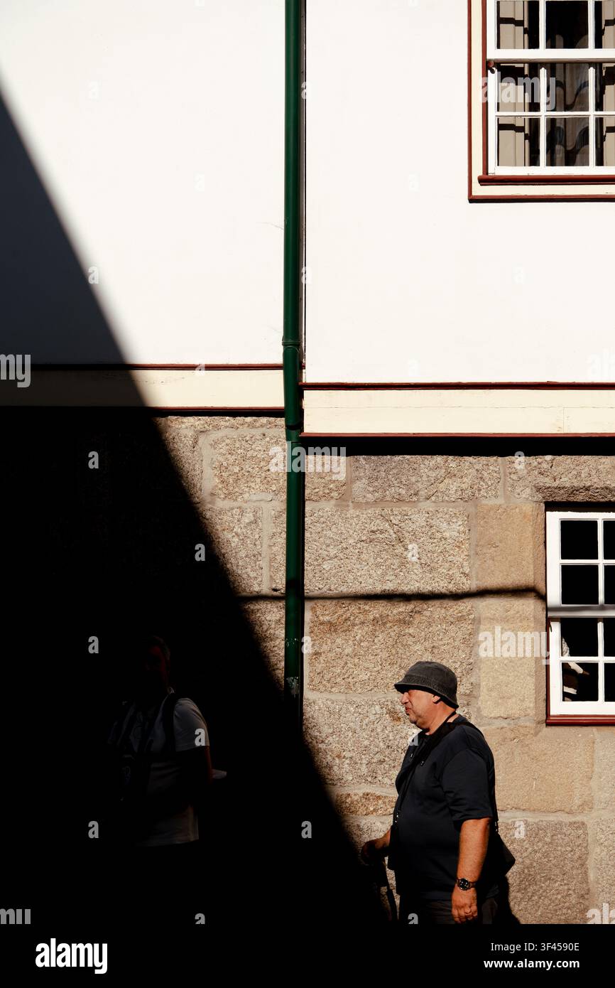 Guimarães, 26. Juli 2026; Mann geht an einem kontrastierenden Schatten und einem grünen Fallrohr an einem alten Stein- und Weißgebäude vorbei. Stockfoto