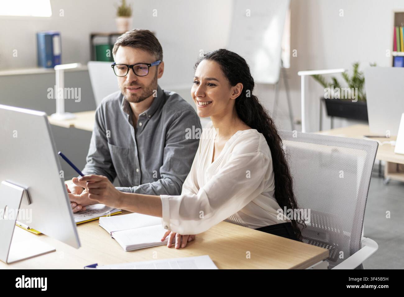 Lächelnde hispanische Frau unterstützt kaukasischen männlichen Kollegen bei der Arbeit am Computer im Büro, Geschäftskonzept Stockfoto