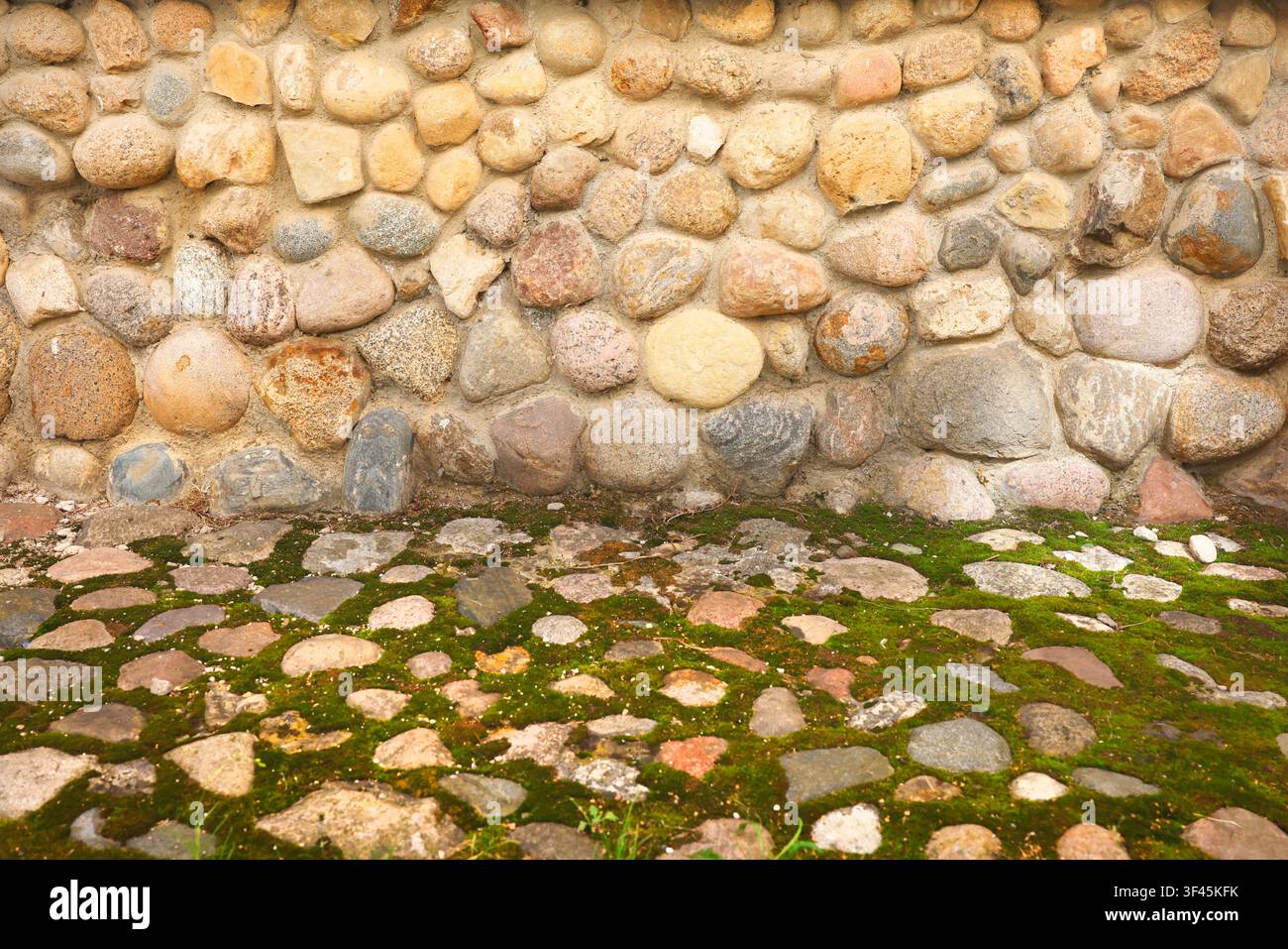 Foto mit Kopfsteinpflaster-Wand, grüner Moos-Bodenhintergrund. Alte rustikale Steinmauerstruktur mit texturierten runden Felsbrocken und altem Kopfsteinpflaster-Boden Stockfoto