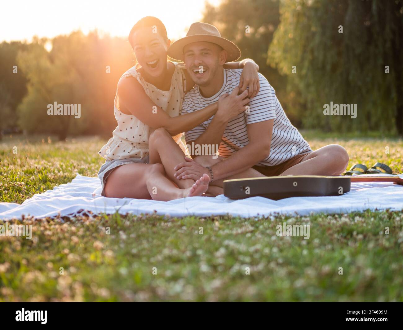 Ein fröhliches multikulturelles Paar sitzt zusammen auf einer Picknickdecke in einem sonnenbeschienenen Park, lacht und umarmt sich während der goldenen Stunde. Stockfoto