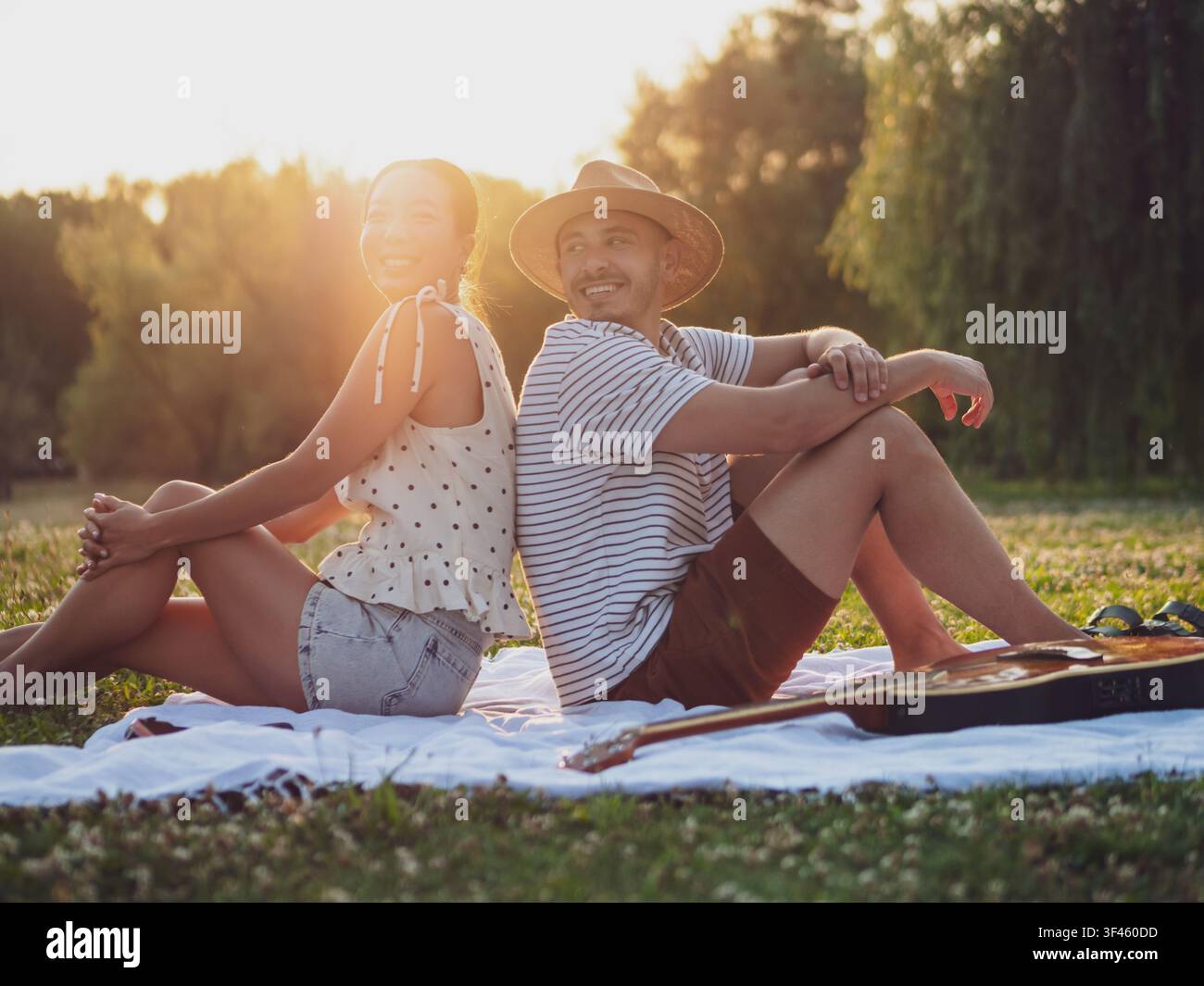 Ein fröhliches Paar teilt einen freudigen Moment auf einer Picknickdecke, umgeben von üppiger Vegetation bei Sonnenuntergang. Stockfoto