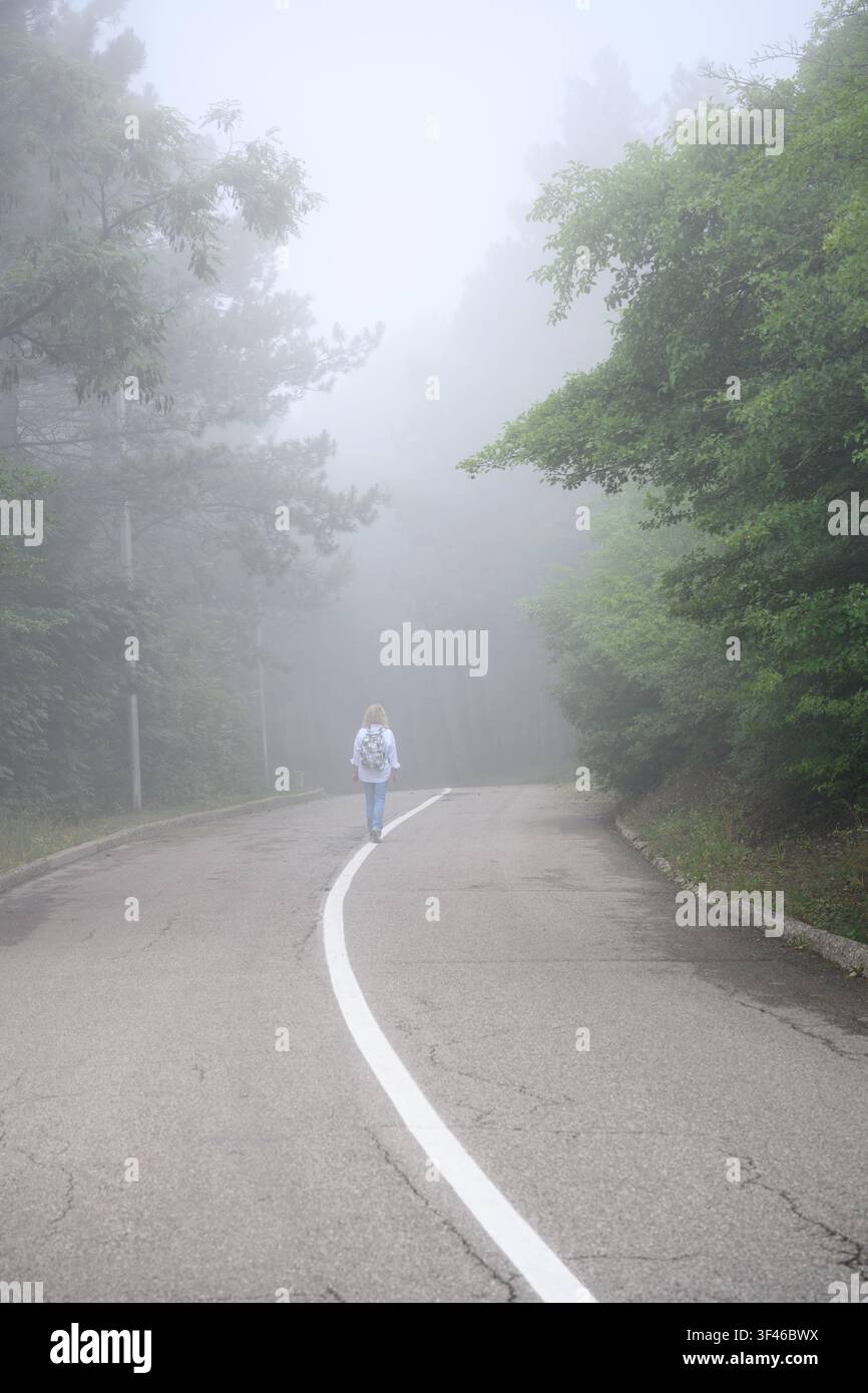 Eine vertikale Aufnahme, die die Rückansicht einer Frau mit einem Rucksack zeigt, die in der Mitte einer asphaltierten Straße geht. Die Straße ist mit einem durchgehenden weißen Streifen markiert Stockfoto