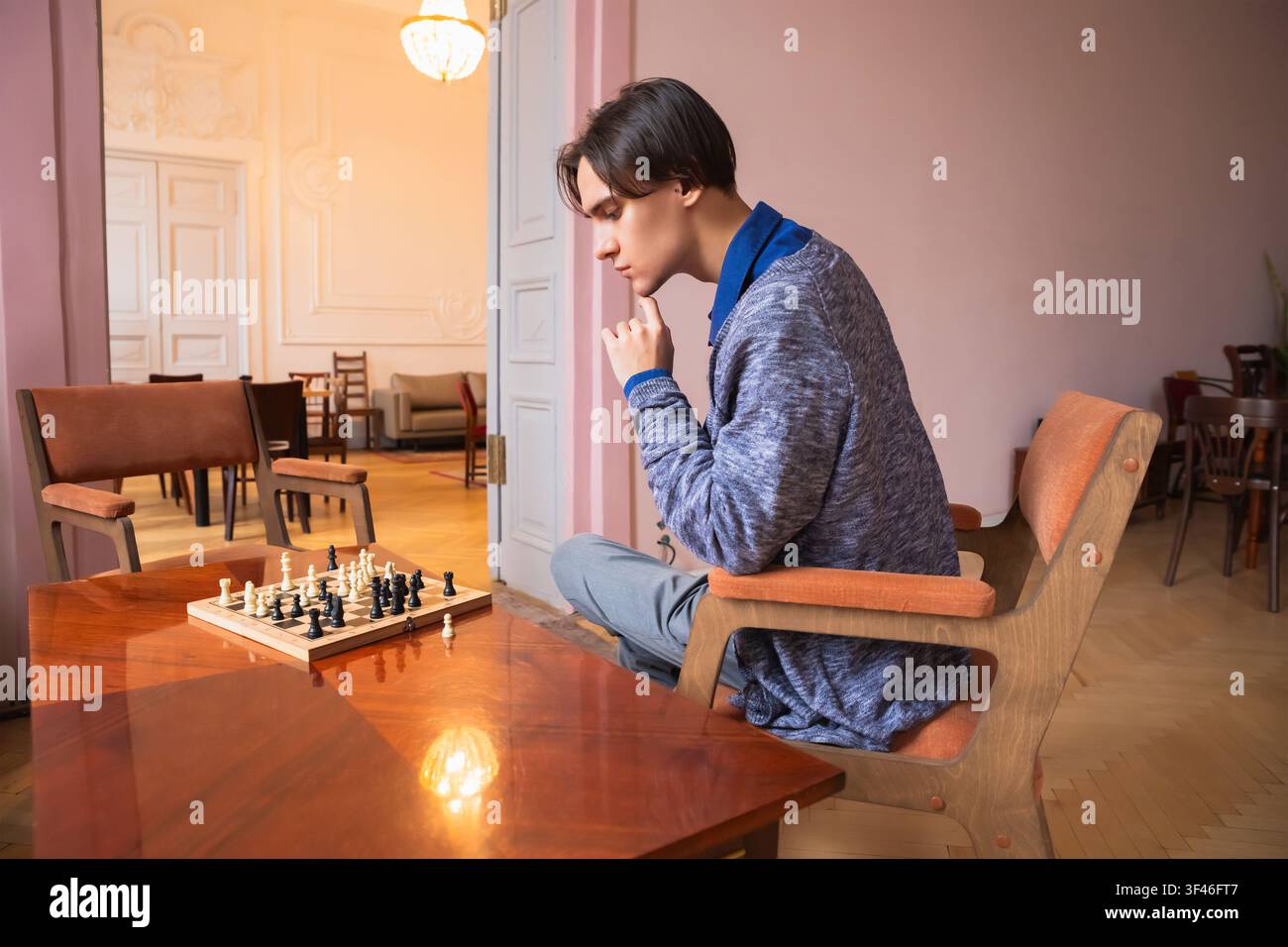Mann spielt Schach, sitzt in einem Sessel und denkt über seinen Zug nach. Konzentrierter junger Student, der während der Freizeit auf das Brettspiel konzentriert ist. Strategie Stockfoto
