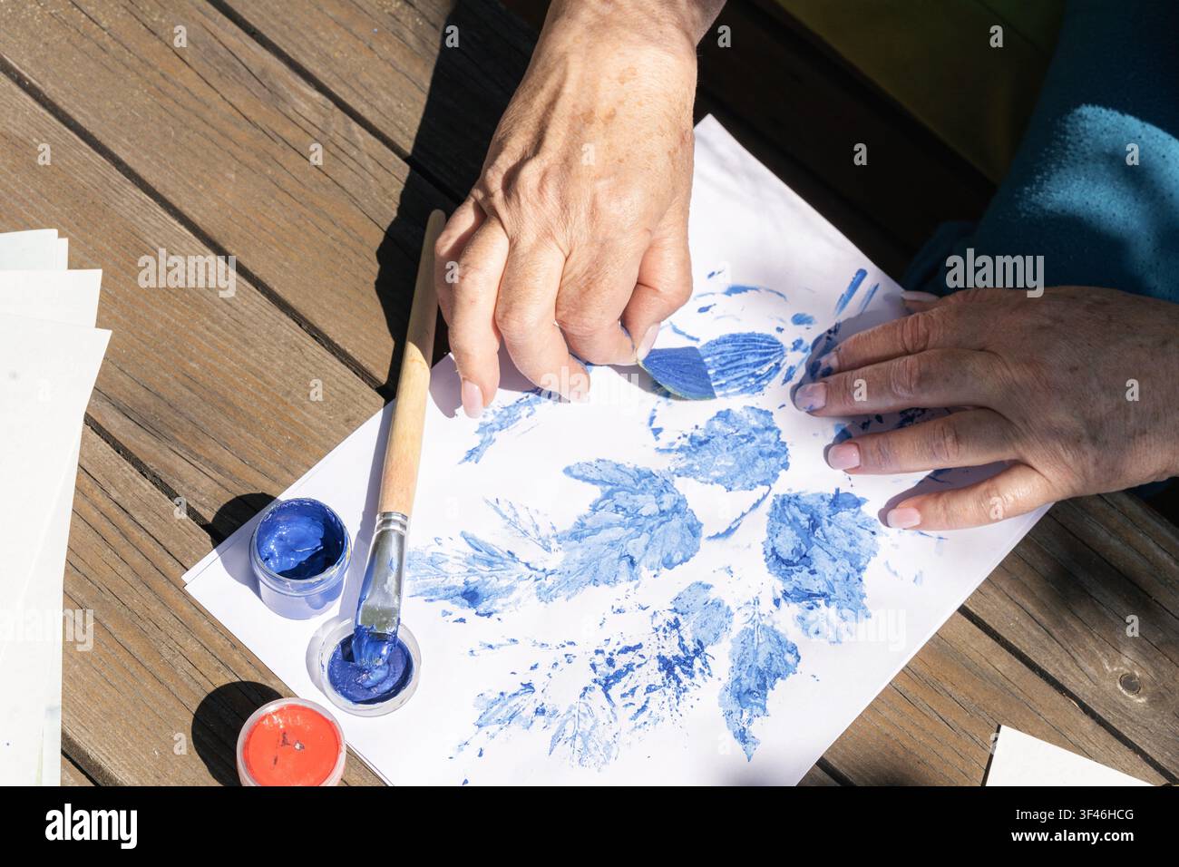 Frauenhände tragen mit dem Pinsel blaue Farbe auf lebende Pflanzenblätter auf und legen sie auf weißes Blatt Papier auf Holztisch, um eine botanische Illustration zu erzeugen Stockfoto