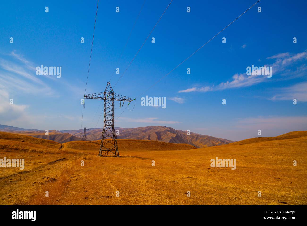 Zwei große Strommasten dominieren eine weite Landschaft mit sanften Hügeln. Trostiges, goldenes Gras deutet auf trockene Bedingungen hin, während ein strahlend blauer Himmel die Stockfoto