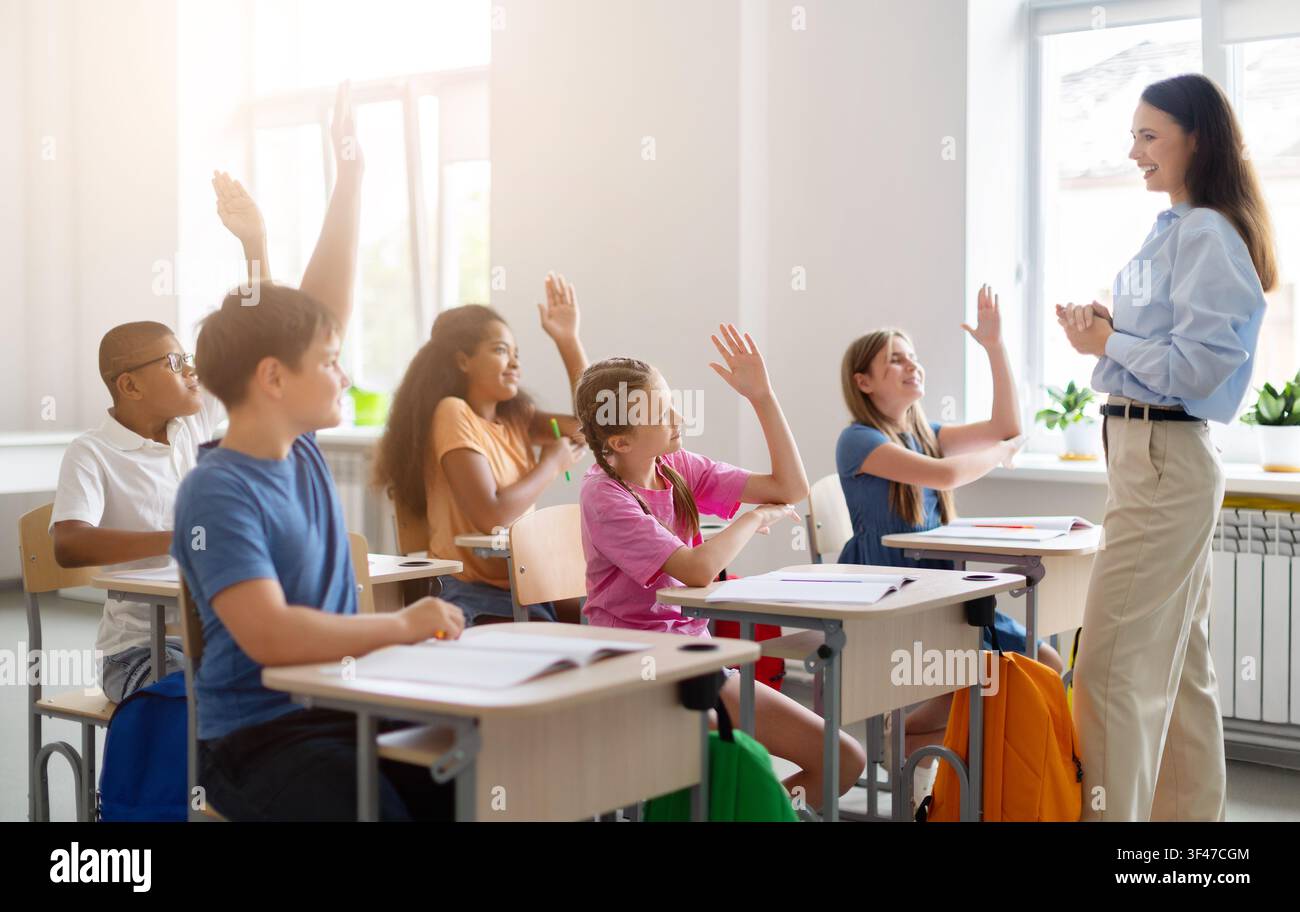 Verschiedene Schulkinder heben die Hände, um einem lächelnden Lehrer während einer interaktiven Unterrichtsstunde zu antworten Stockfoto