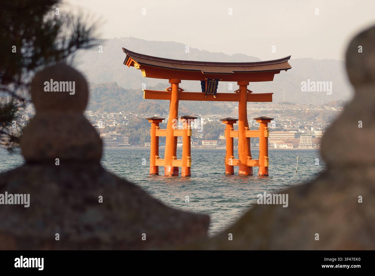 Traditionelle Steinmauer im Vordergrund, die einen entfernten Blick auf das große schwimmende rote Torii-Tor über dem Ozeanwasser freigibt, Hiroshima, Japan. Stockfoto