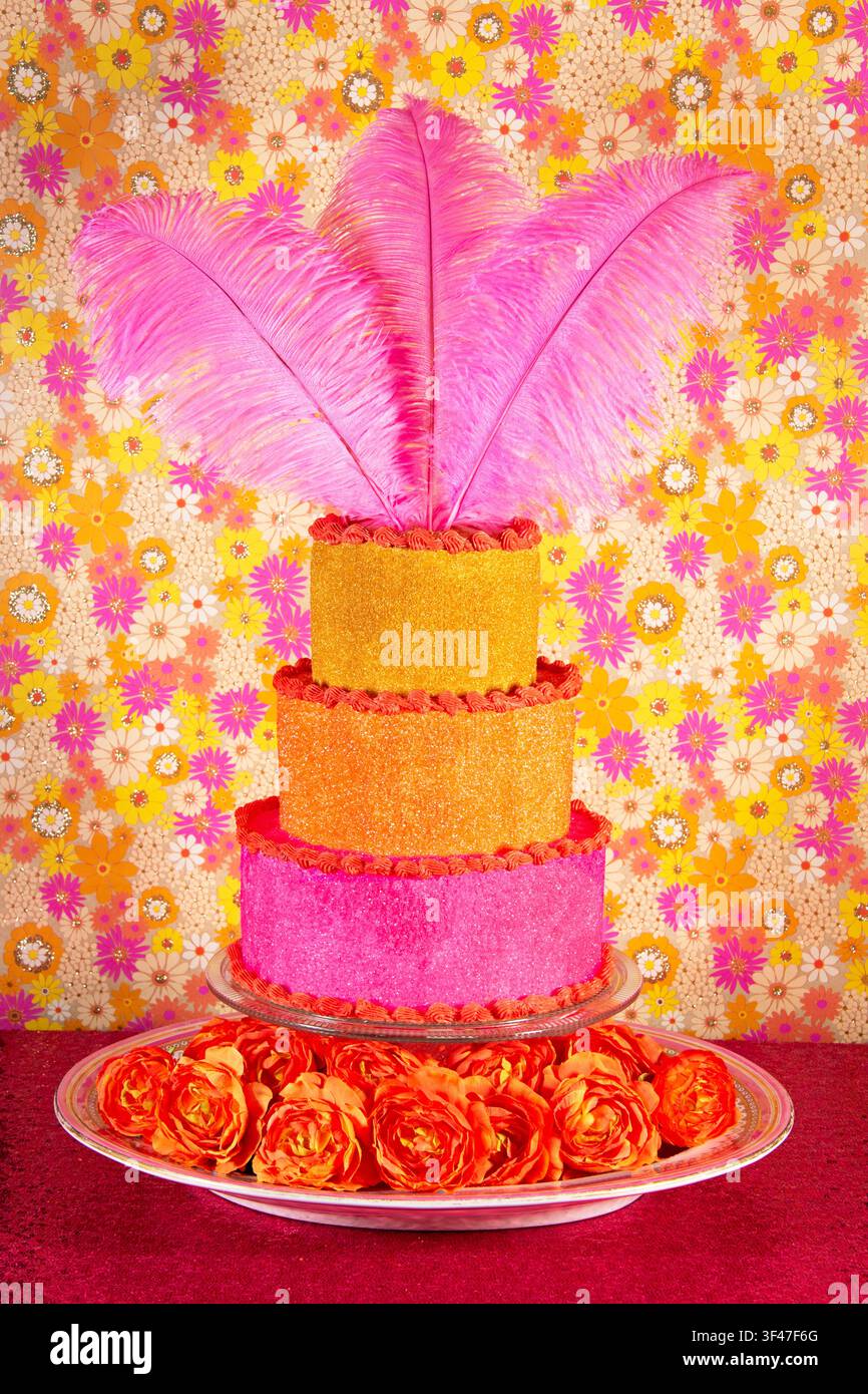 Drei Stockwerke Kuchen dekoriert mit rosa, orange und goldenem Streuzucker, umrandet mit leuchtend rosa Kaugummibällchen; Teller gefüllt mit Blumen; Pop-Art-Gänseblümchen-Tapete. Stockfoto