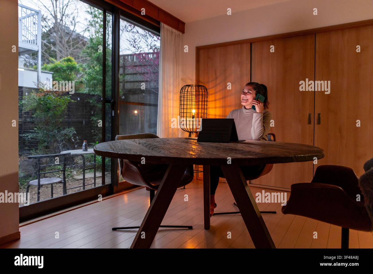 Eine Frau arbeitet von zu Hause aus, sitzt an einem Holztisch, führt ein Telefongespräch und nutzt dabei ein Tablet. Warme Beleuchtung schafft eine gemütliche Atmosphäre im r Stockfoto