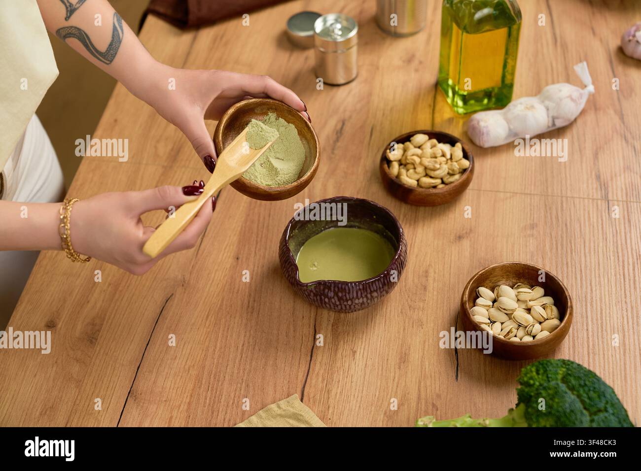Hände mischen lebendigen Matcha-Puder mit Nüssen und Ölen in einer gemütlichen Kücheneinrichtung. Stockfoto