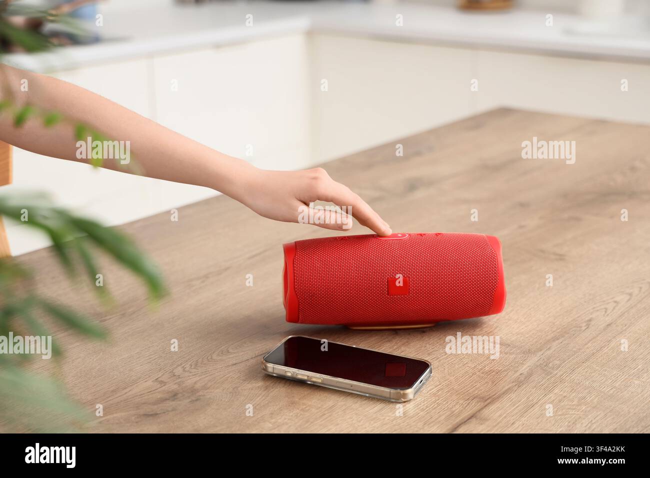 Person, die einen roten tragbaren Bluetooth-Lautsprecher neben einem Smartphone auf einem Holztisch einschaltet, im Hintergrund ist eine moderne Küche mit weißen Schränken zu sehen Stockfoto