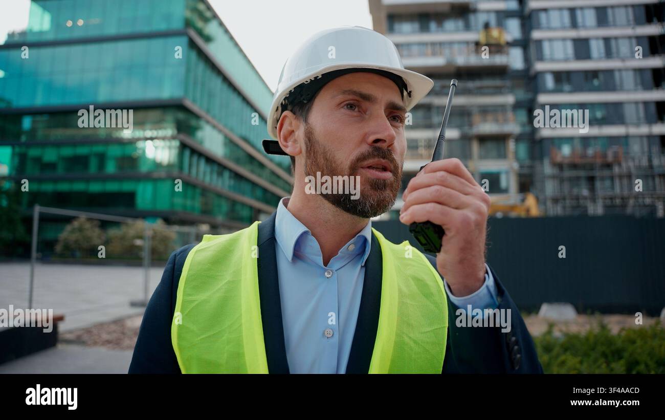 Moving Shot Kaukasischer Mann Bauingenieur in Hardhat Talk Remote Radio Outdoor Urban Building Architekt Bauunternehmer mit Helm Sicherheitsweste Stockfoto