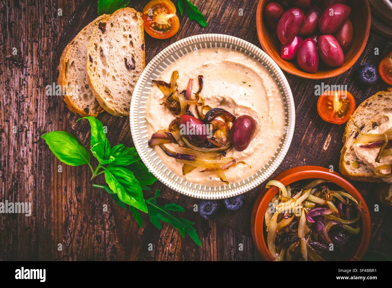 Köstlicher Hummus mit karamellisierten Zwiebeln, Oliven und frischem Brot auf einem Holztisch Stockfoto