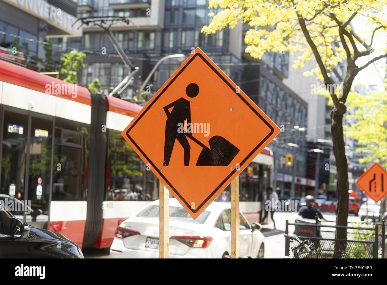 Ein helles orangefarbenes Baustellenschild steht auffällig auf einer belebten Straße in Toronto, mit einer modernen rot-weißen Straßenbahn, Autos und Fußgängern im Stockfoto