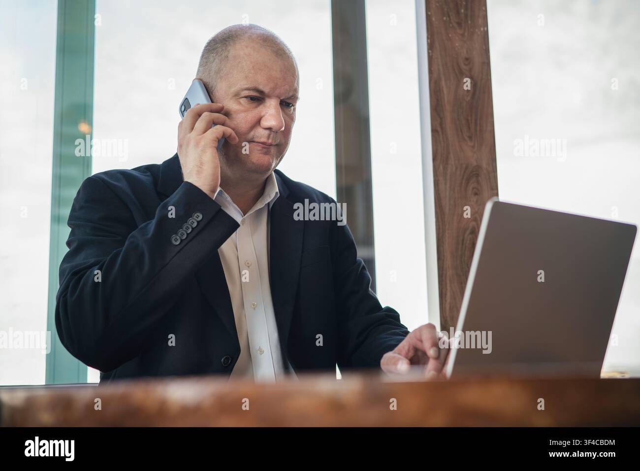 Ernster reifer Geschäftsmann, der am Smartphone spricht, während er an einem Laptop in einem modernen Arbeitsbereich arbeitet. Geschäftskommunikation, Multitasking und Fernarbeit con Stockfoto