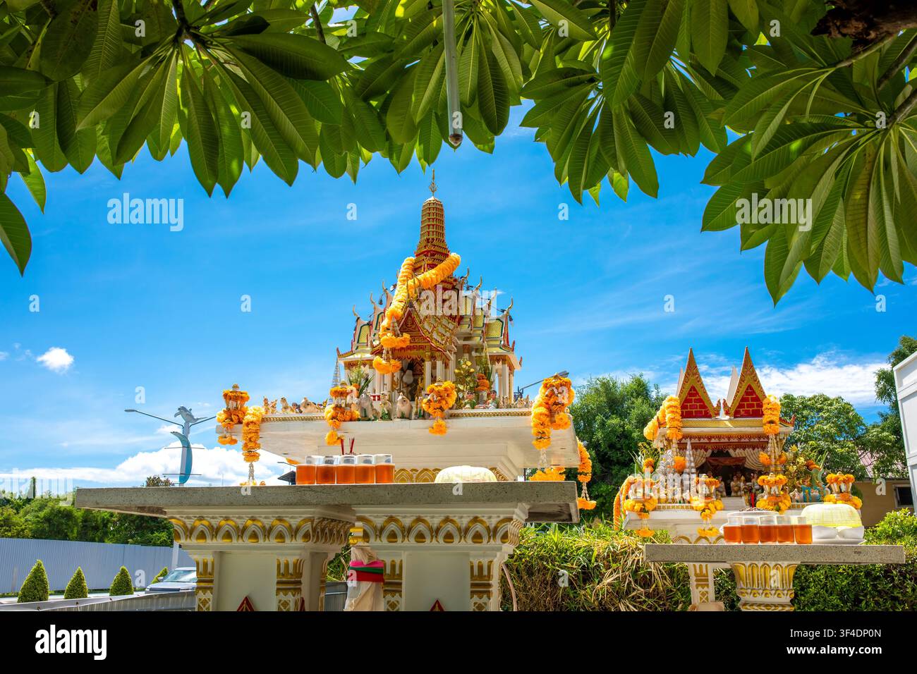 Traditioneller thailändischer Geisterhaus-Schrein, geschmückt mit Blumengirlanden, eingerahmt von grünen tropischen Baumblättern unter blauem Himmel Stockfoto