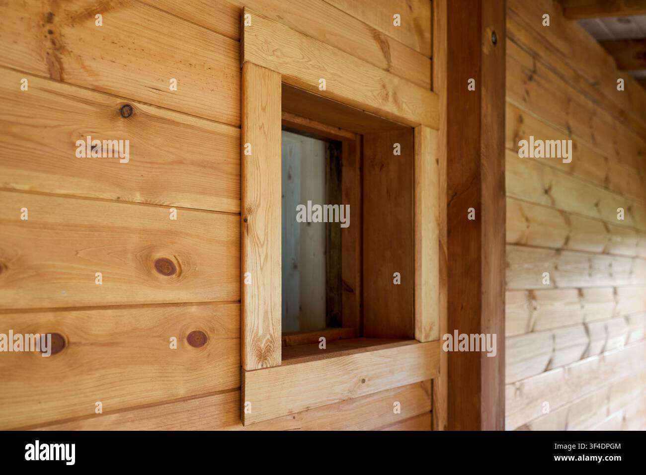 Nahaufnahme eines kleinen quadratischen Glasfensters, das in eine rustikale Holzwand aus gezapften Kiefernholzplanken mit heller Naturlackierung integriert ist, das ein w Stockfoto