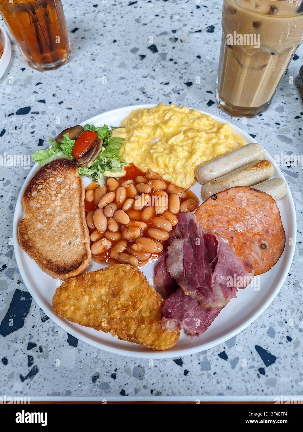 Nahaufnahme eines traditionellen englischen Vollfrühstückstellers mit Rührei, Baked Beans, Würstchen, Toast und einem Hash Brown auf einem Terrazzotisch. Stockfoto