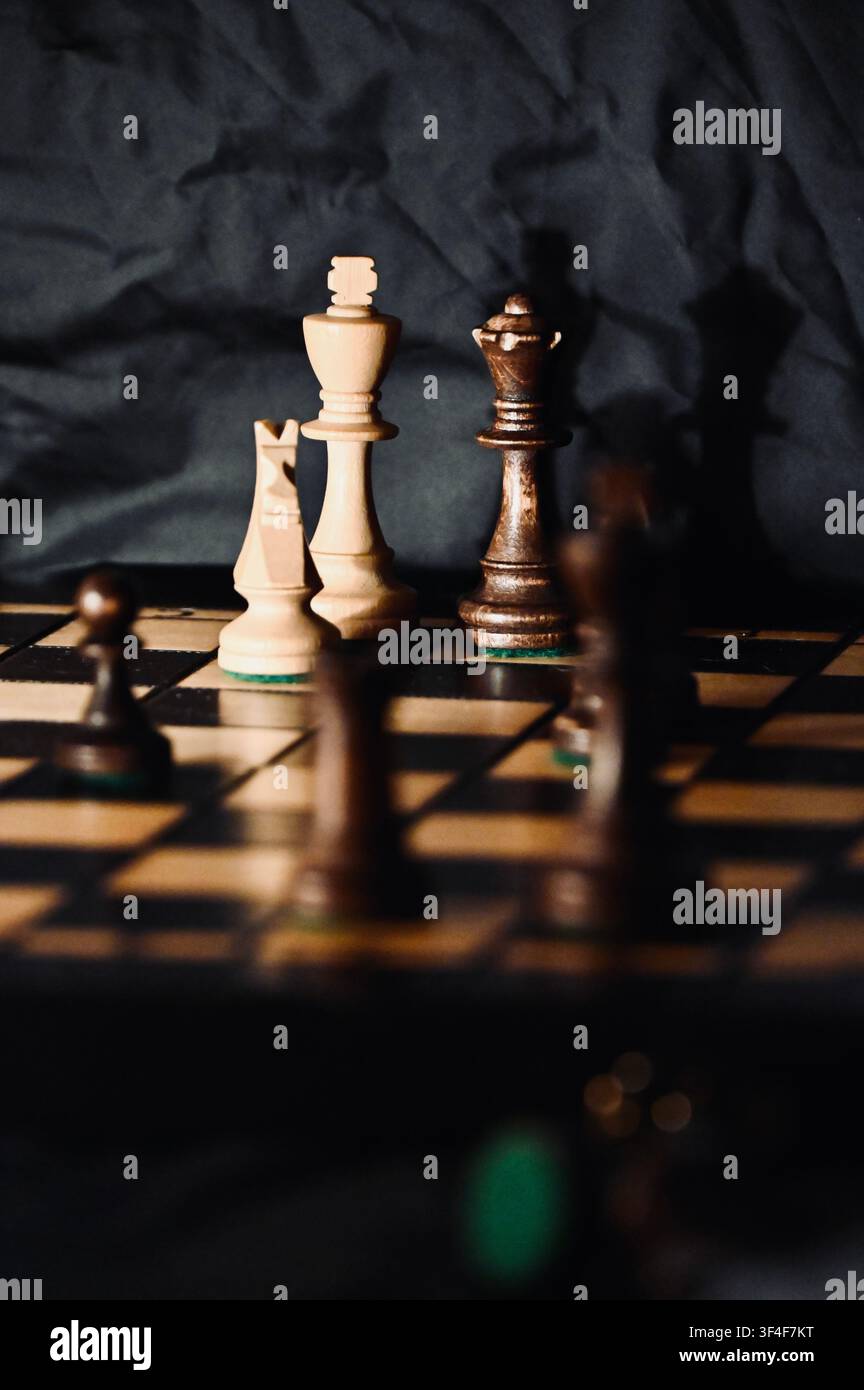 Ein stimmungsvolles, gedämpftes Foto eines Holzschachbretts mit Fokus auf die Hauptfiguren, einschließlich des weißen Königs, des Springers und der schwarzen Dame. Tiefe Schatten, atmosphärische dramatische Beleuchtung und ein dunkler, zerknitterter Hintergrund erzeugen ein starkes Gefühl von Strategie, Wettbewerb, Spannung und Intellekt. Perfekt für Konzepte, die sich auf Geschäftsstrategie, Entscheidungsfindung, Taktik und klassische Tischspiele beziehen. Stockfoto