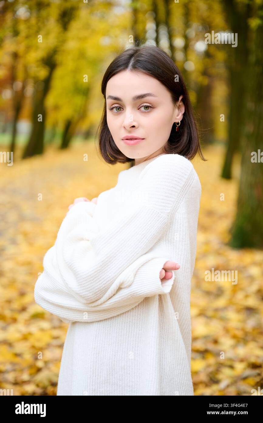Porträt einer jungen Frau, die in einen weichen weißen Pullover gehüllt ist und in einem Herbstpark mit gelben Blättern und verschwommenen Bäumen im Hintergrund posiert. Perfekt für Mode, gemütliches Lifestyle, Schönheit, saisonale Werbung und Anzeigen Stockfoto