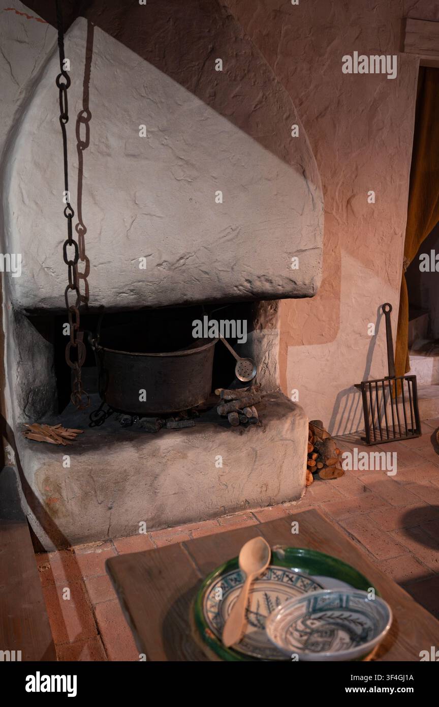 Rustikal traditionelles Kücheninterieur mit einem Stein-Kamin mit einem Kochtopf über offenem Feuer und Keramikgeschirr auf einem Holztisch im Vordergrund. Stockfoto