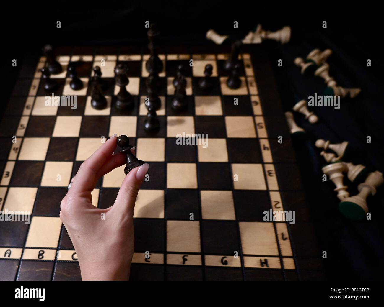 Eine obere, stimmungsvolle Ansicht der Hand eines Spielers, die eine schwarze Schachfigur hält und eine Entscheidung auf einem hölzernen Schachbrett trifft. Dunkle und helle Schachfiguren sind in einer taktischen Position angeordnet, wobei eroberte Figuren neben dem Brett ruhen, was Geschäftsplanung, Führung, Entscheidungsfindung und Strategie symbolisiert. Stockfoto