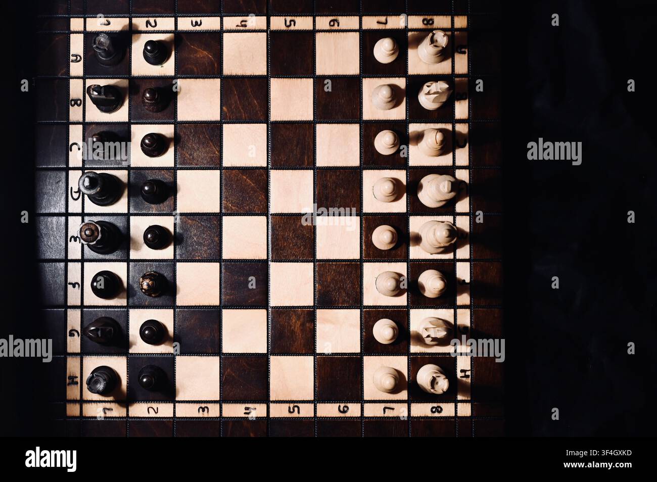 Hochwinklige Flachlay-Ansicht eines Holzschachbretts mit allen Figuren in Startposition angeordnet. Der dramatische Kontrast zwischen hellen und dunklen Tönen mit subtilen Schatten symbolisiert strategische Planung, Geschäftskonkurrenz, taktisches Denken, Entscheidungsfindung und intellektuelle Herausforderung. Stockfoto
