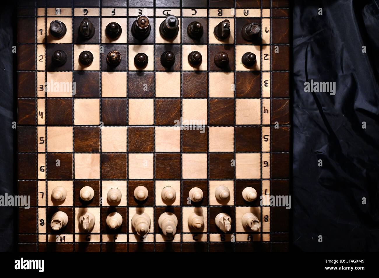 Hochwinklige Flachlay-Ansicht eines Holzschachbretts mit allen Figuren in Startposition angeordnet. Der dramatische Kontrast zwischen hellen und dunklen Tönen mit subtilen Schatten symbolisiert strategische Planung, Geschäftskonkurrenz, taktisches Denken, Entscheidungsfindung und intellektuelle Herausforderung. Stockfoto