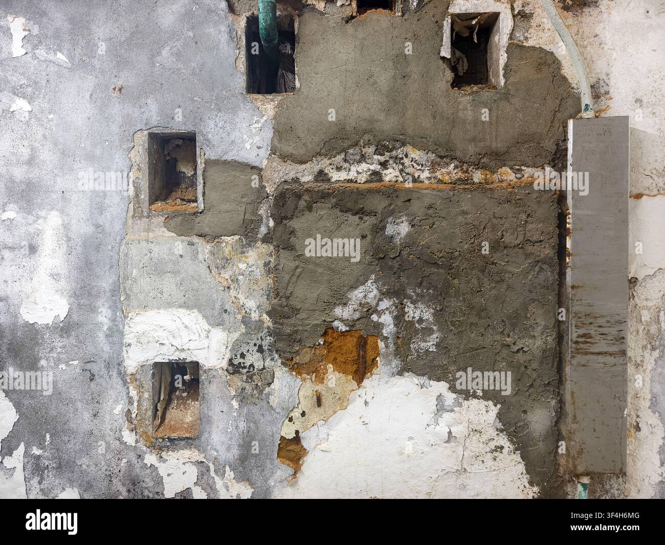 beschädigte, unebene Wand, die grobe Mauerreparaturen mit freiliegenden Hohlräumen, Schichten von altem Putz, abblätternder Farbe und frischen Zementflecken erfährt Stockfoto