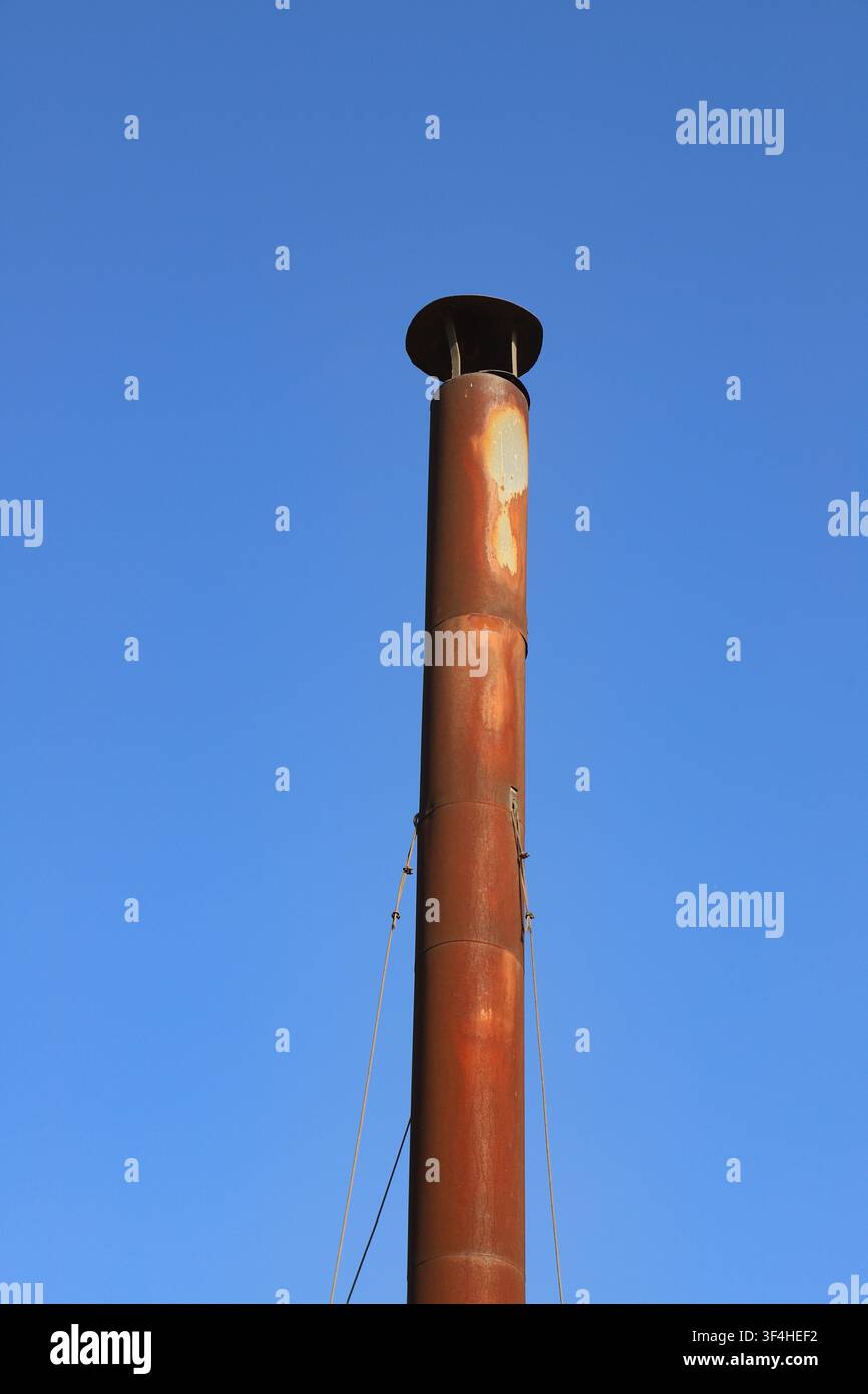 Diese Nahaufnahme zeigt einen hohen, verrosteten Metallschornstein auf einer verlassenen Industriebrache im Freien. Der orange-braune Rost bedeckt den Zylinder, und Stockfoto