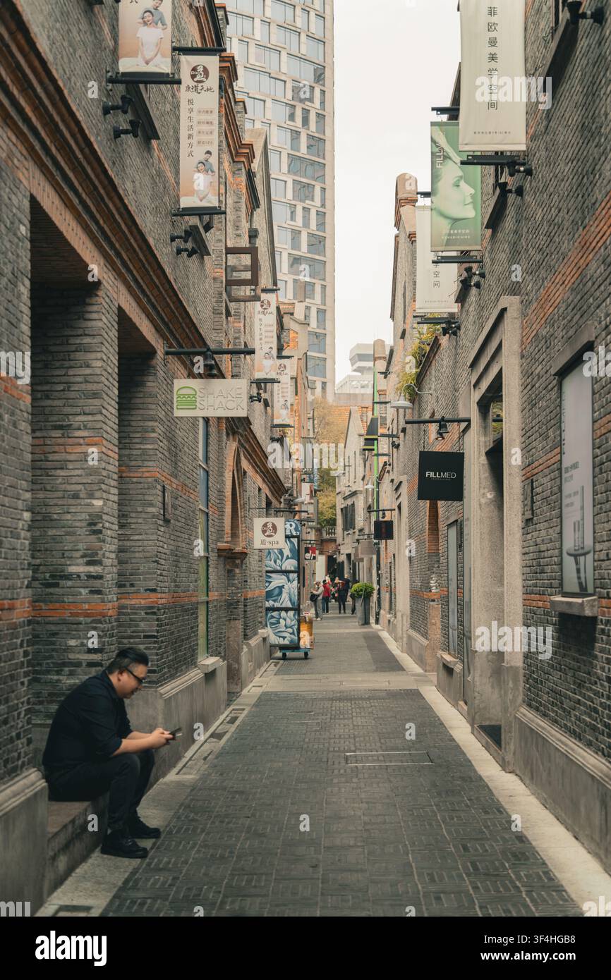 Mann sitzt auf der Straße und verwendet ein Smartphone in einer engen historischen Backstein-Gasse, Xintiandi, Shanghai Stockfoto
