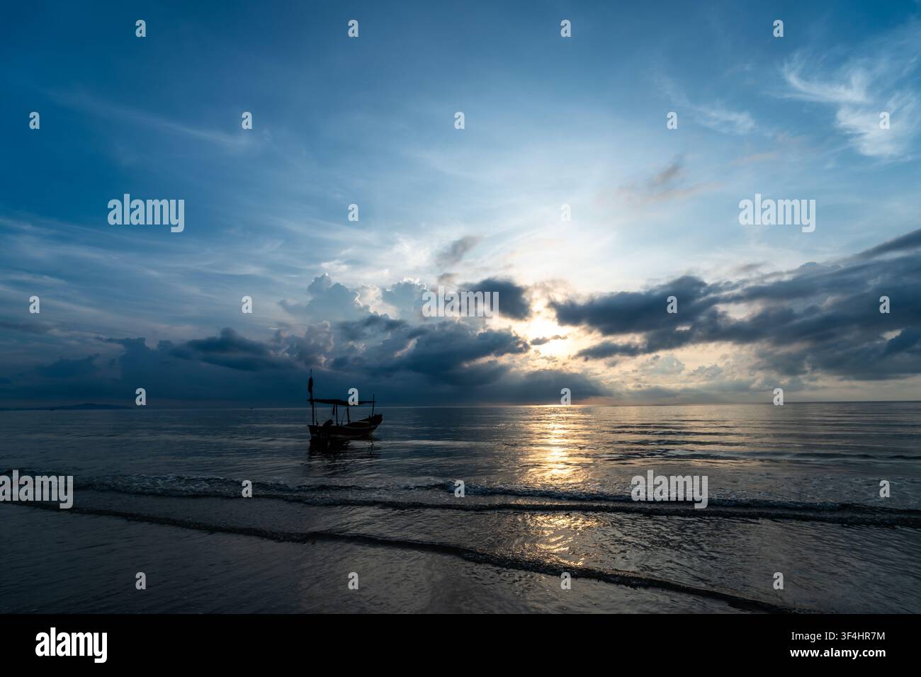 Ein traditionelles Langrumpf-Fischerboot ruht auf dem ruhigen tropischen Meer bei Sonnenaufgang, als Silhouette vor einem dramatischen Himmel, durch den goldenes Licht bricht Stockfoto
