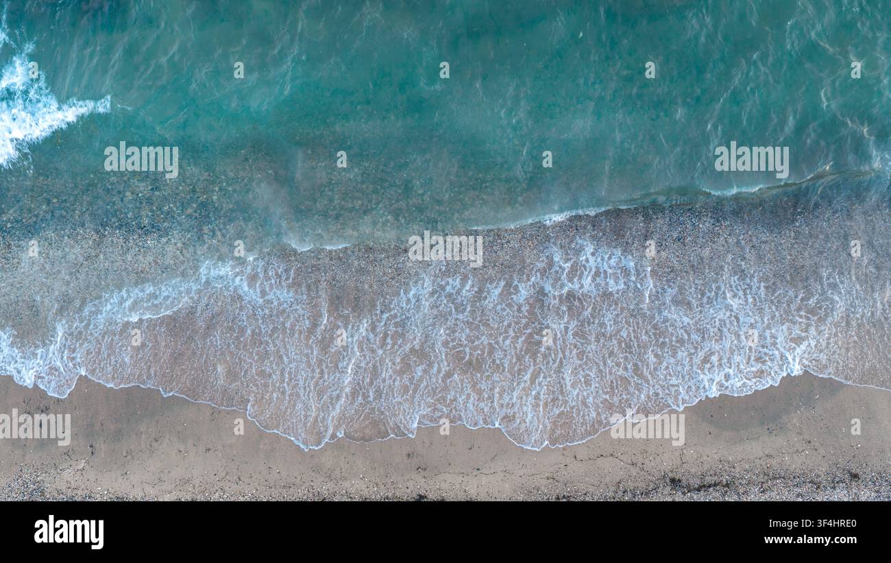 Top-Down-Drohnenaufnahme fängt schaumige Wellen ein, die über klares blaues Wasser auf einen breiten Kiesstrand in Nea Moudania, Griechenland, rollen. Stockfoto