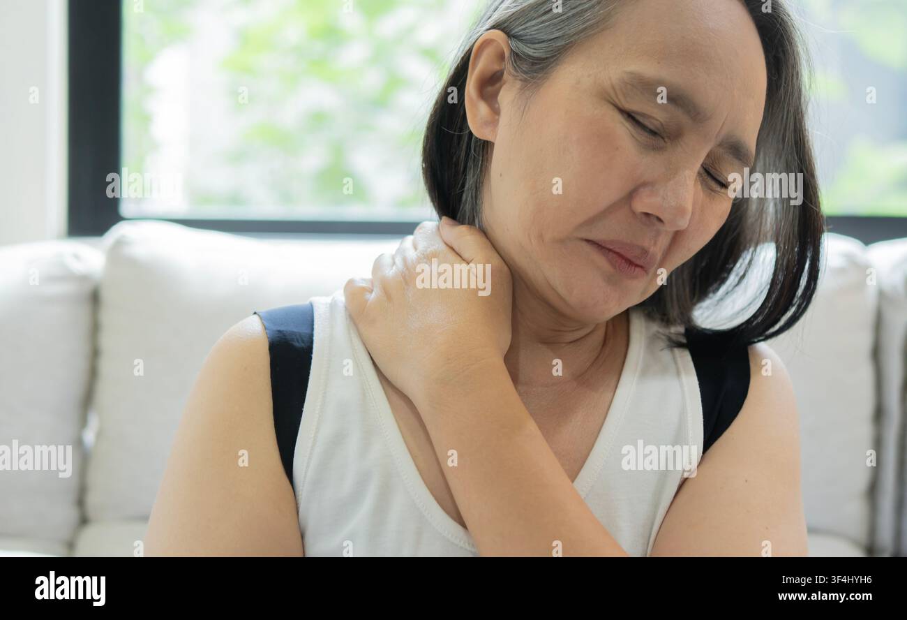 Asiatische reife Frau berührt ihren Hals wegen Rückenschmerzen und streckt die Arme. Frau massiert ihren Rücken bei Schmerzen und schließt die Augen, um sich zu Hause zu entspannen. Stockfoto