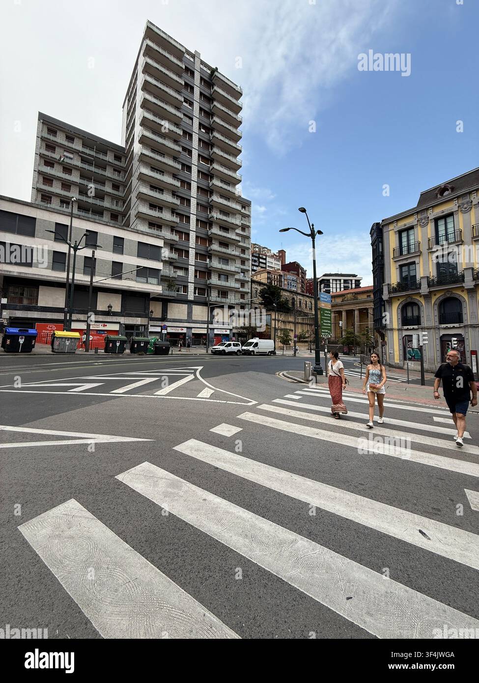 Fußgänger überqueren eine breite städtische Kreuzung, umgeben von gemischten historischen und modernen Gebäuden im Zentrum von Bilbao, Spanien. - Smartphone-aufgenommenes Stockfoto