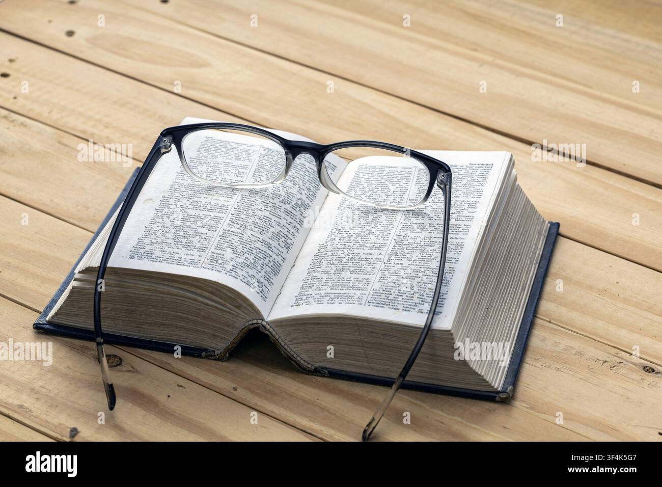 Offenes Wörterbuch mit schwarz gerahmter Lesebrille auf den Seiten, die auf einem Holztisch ruhen, Konzept der Kurzsichtigkeit, Bildung und Studium Stockfoto