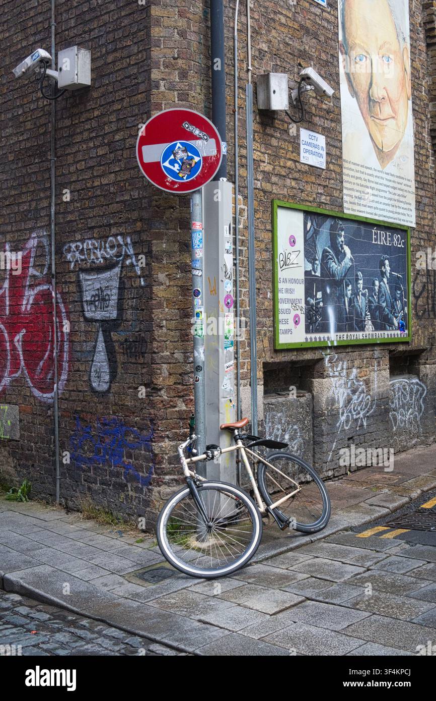 Fahrrad, das an einem Pfosten an einer Kopfsteinpflaster-Eckstraße mit Graffiti befestigt ist. ein Halteverbotsschild und Sicherheitskameras in Dublin, Irland. Stockfoto