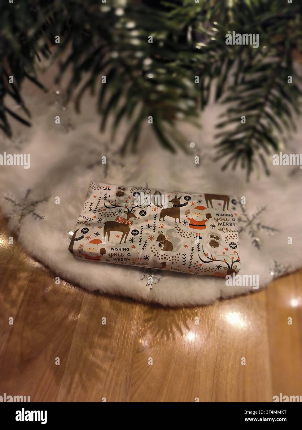 Ein wunderschön verpacktes Weihnachtsgeschenk mit festlichen Tiermustern, das unter einem Weihnachtsbaum auf einem weißen Kunstpelzrock platziert ist. - Smartphone-aufgenommenes Stockfoto