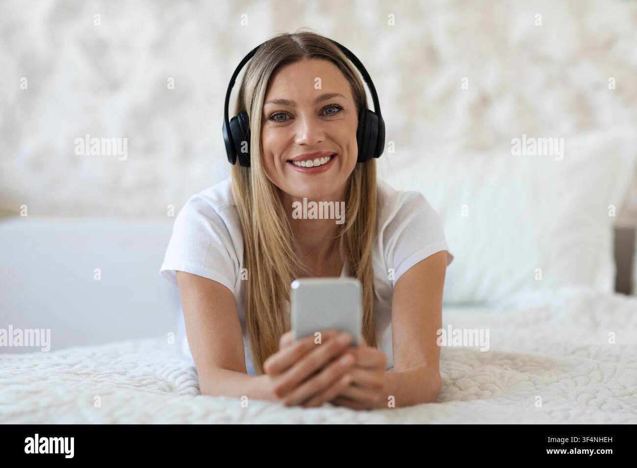 Zufriedene junge Frau hört Musik mit Kopfhörern im Bett, entspannt zu Hause Stockfoto